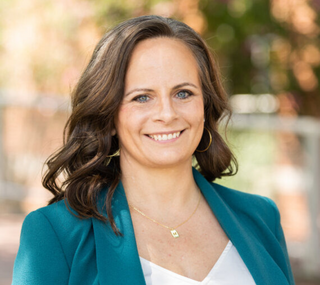 Eliza McCoy in a aquamarine suit jacket and white blouse smiling at the camera in front of a blurred background